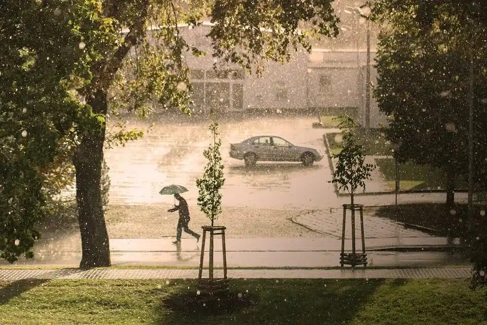 Photo d'un paysage pluvieux figurant un personnage courant sous la pluie avec un parapluie. En arrière plan, une voiture.